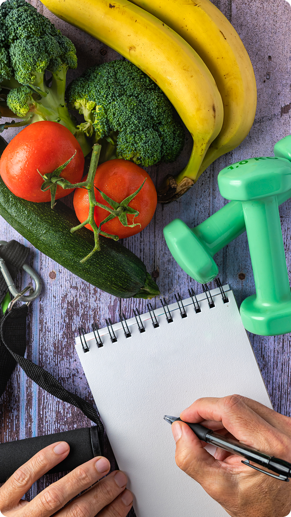 Notebook, fruits, vegetables, and dumbbells on table.